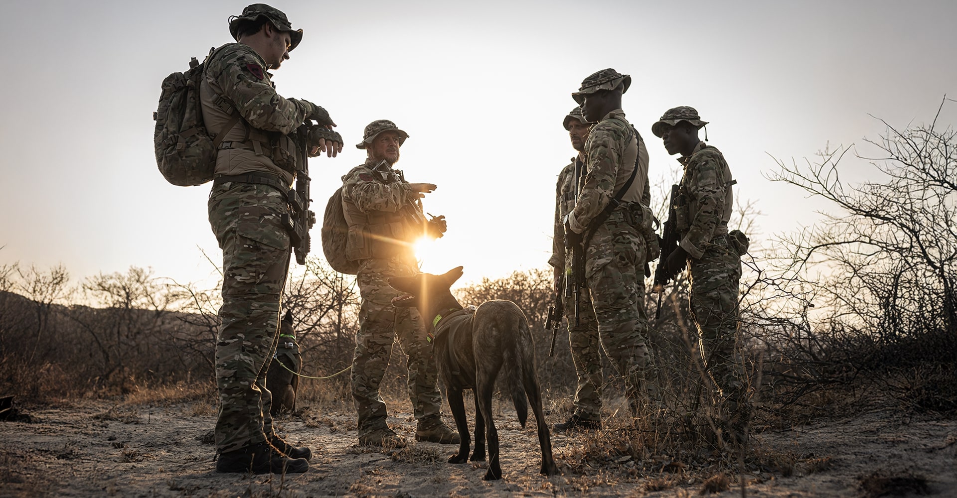 Five operators, including park rangers and K9 handlers, stand in a circle with a K9 dog at the center, discussing tactics in the early morning sun.