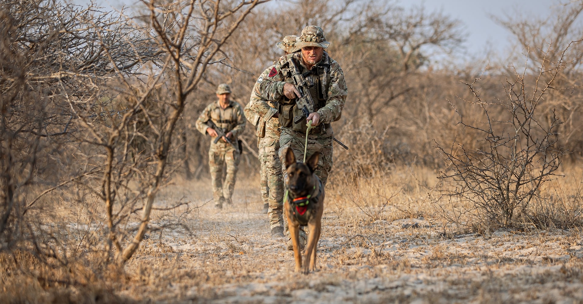 A K9 on a leash, following its handler’s lead as they track down poachers through Africa’s rugged terrain. In the background, another handler and dog are also in pursuit.