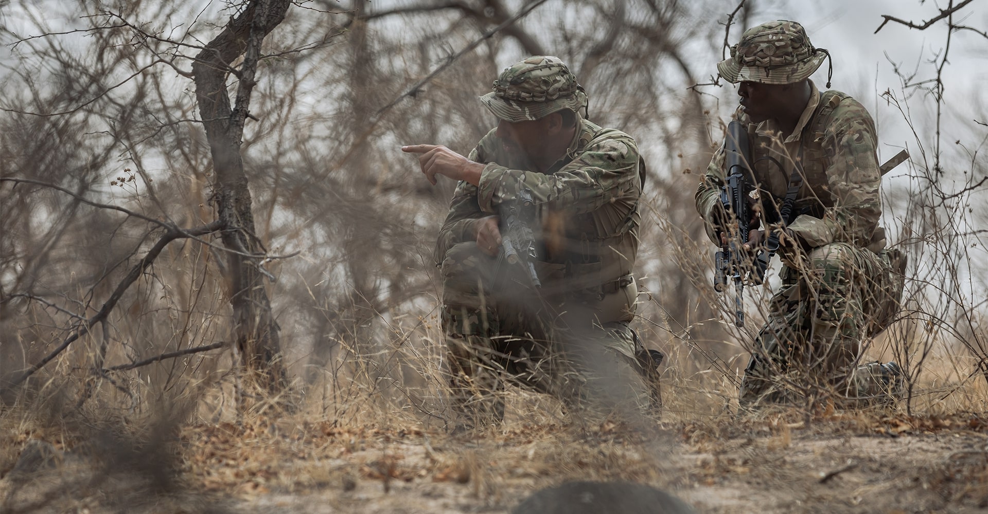 Two park rangers concealed in the bush, one gesturing toward a suspected area of activity.