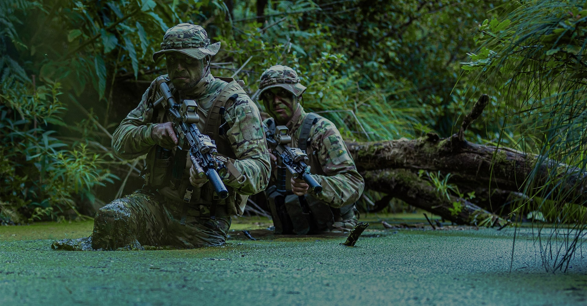 Two operators in full military BDU wading through murky jungle water, surrounded by dense green vegetation.