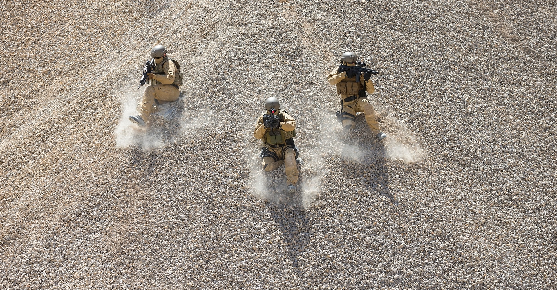 Three operators sliding down a rocky hill on their knees in a quarry-like environment.