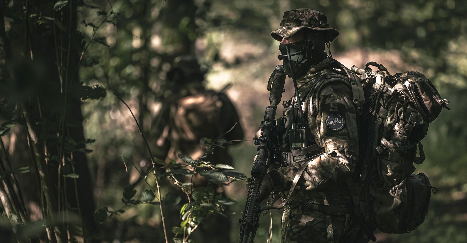 A fully camouflaged operator stands in the woods wearing a MultiCam battle dress uniform.
