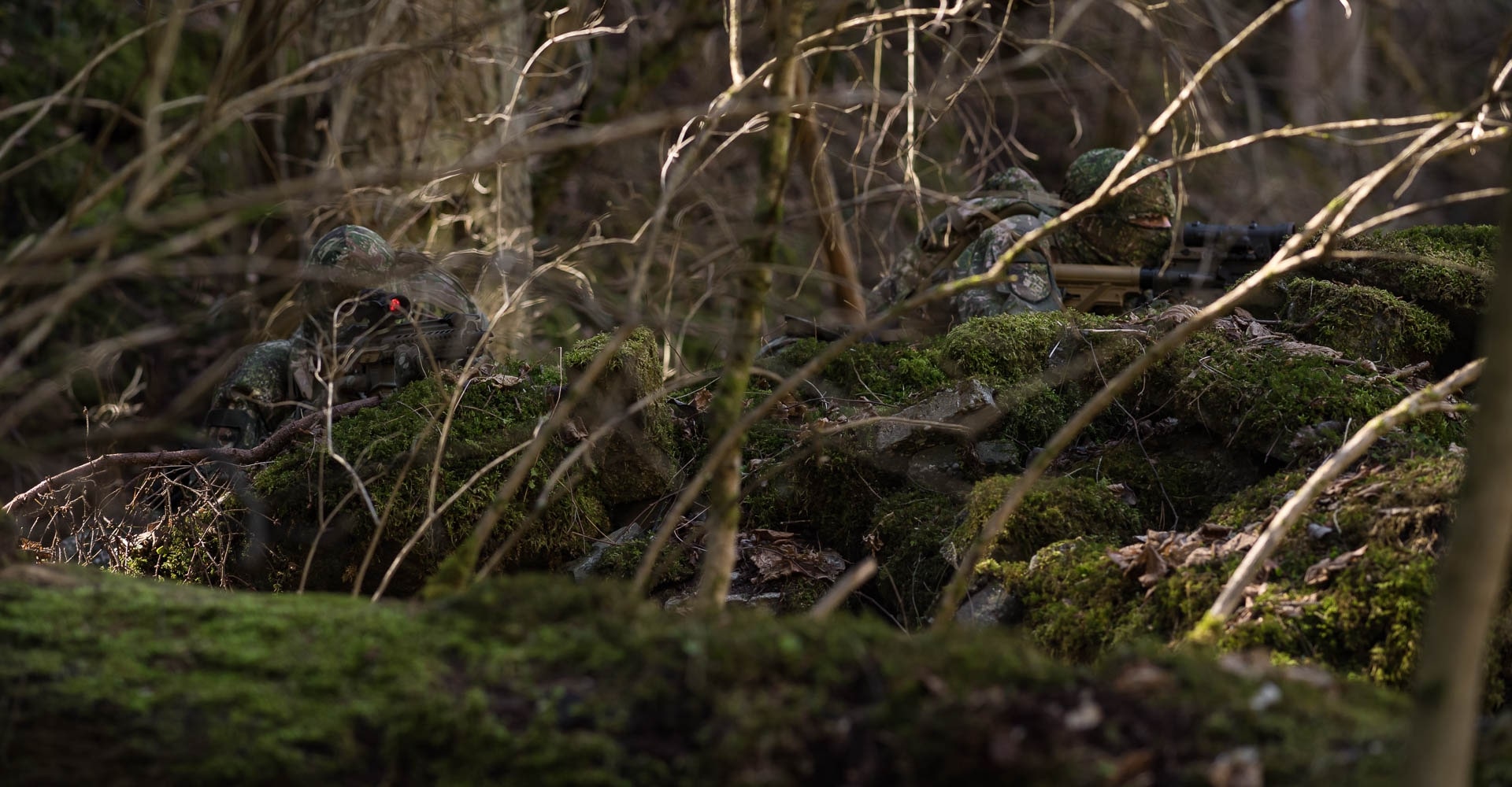 Two operators in full camouflage take cover behind fallen tree logs.