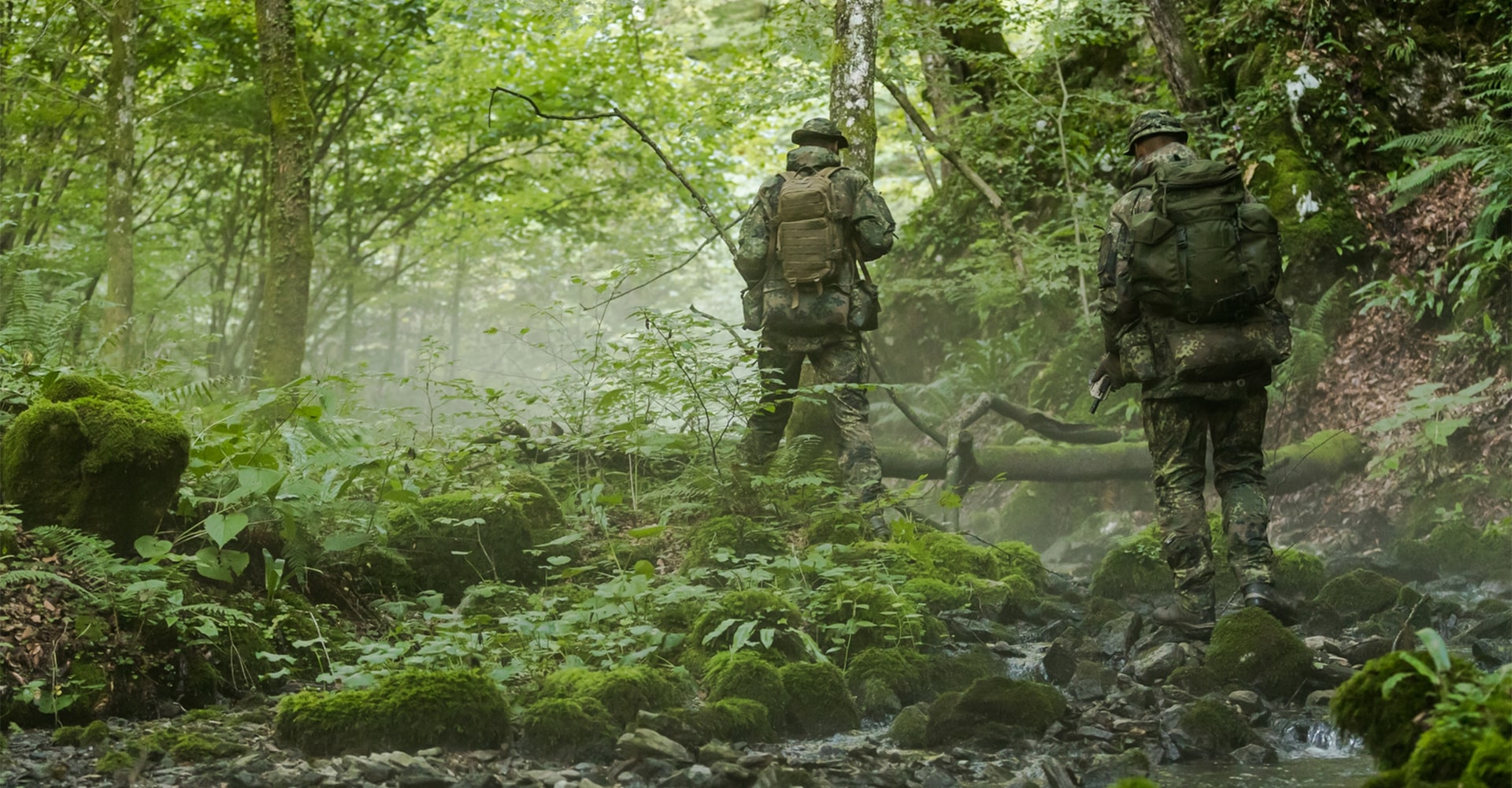 Two operators move through wooded terrain alongside a shallow stream, blending seamlessly into the environment with their camouflage gear.