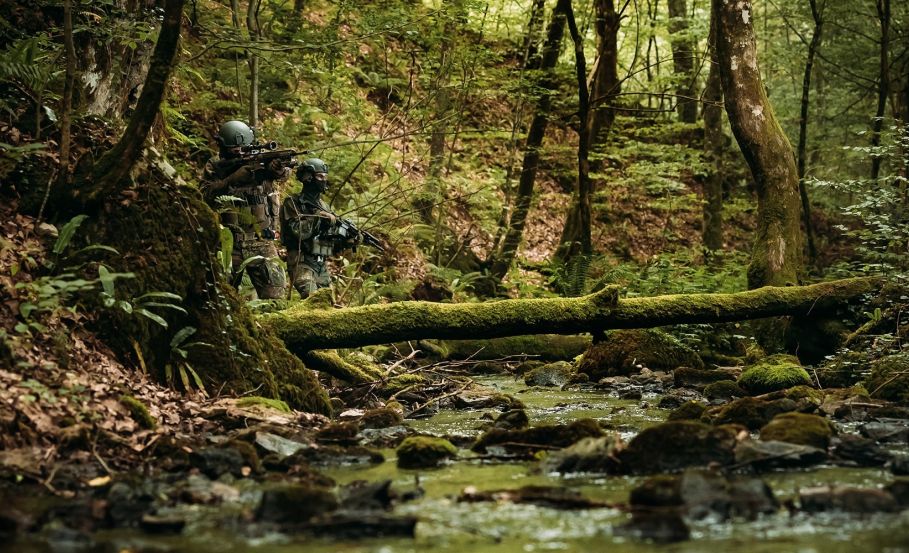 Two operators in wooded terrain with a small stream nearby, both in full camouflage gear—one aiming into the distance, the other scanning the surroundings. Camouflage Patterns: The Art and Science Behind Tactical Concealment