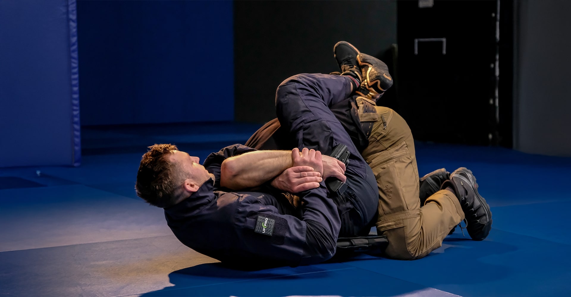 The fight transitions to the ground, with the practitioner securing the opponent’s shoulder and head, grabbing the wrist to control the gun, while controlling their waist using leg positioning.