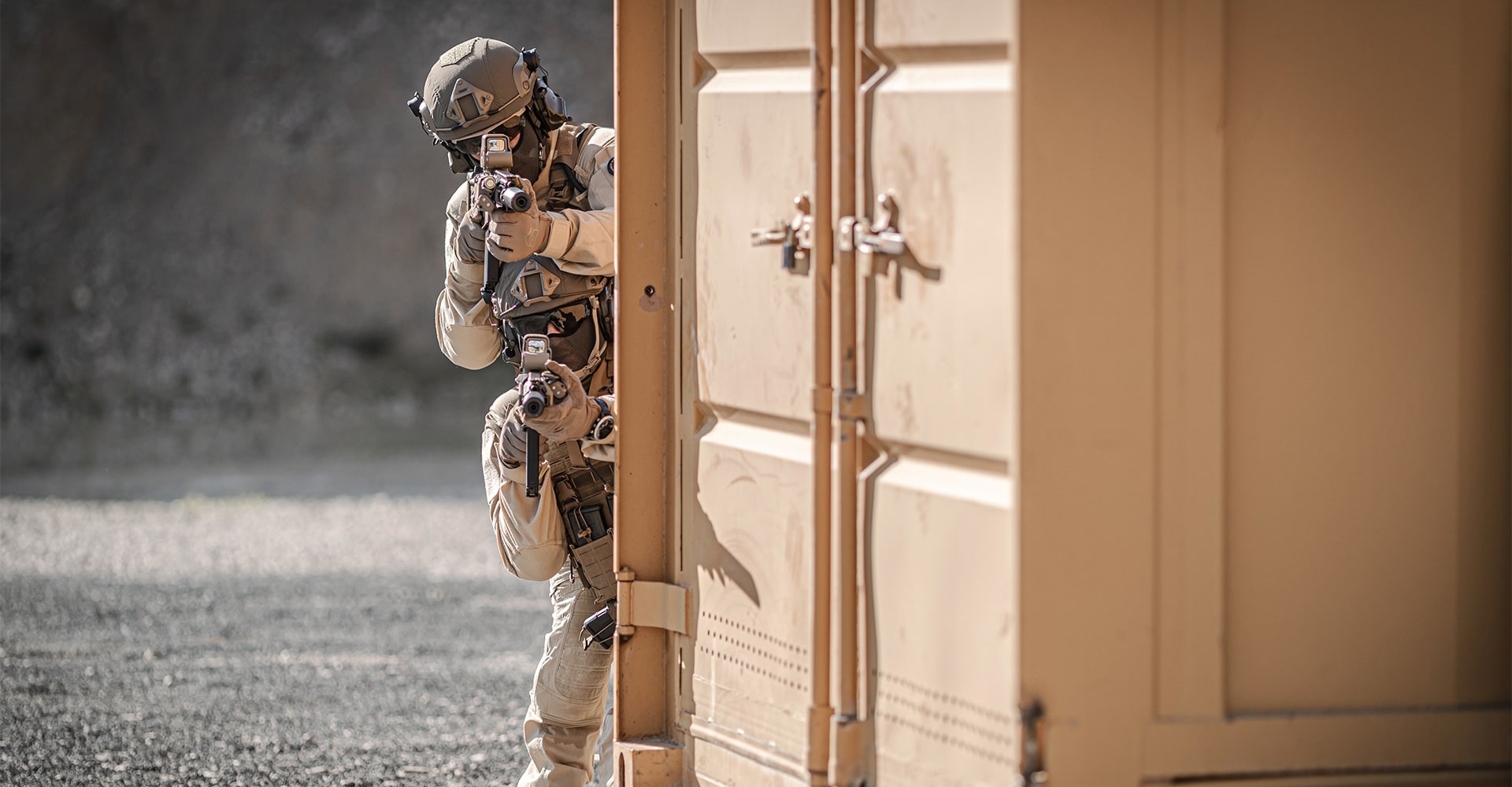 Operator taking cover behind a container in the desert, aiming a weapon toward the camera.