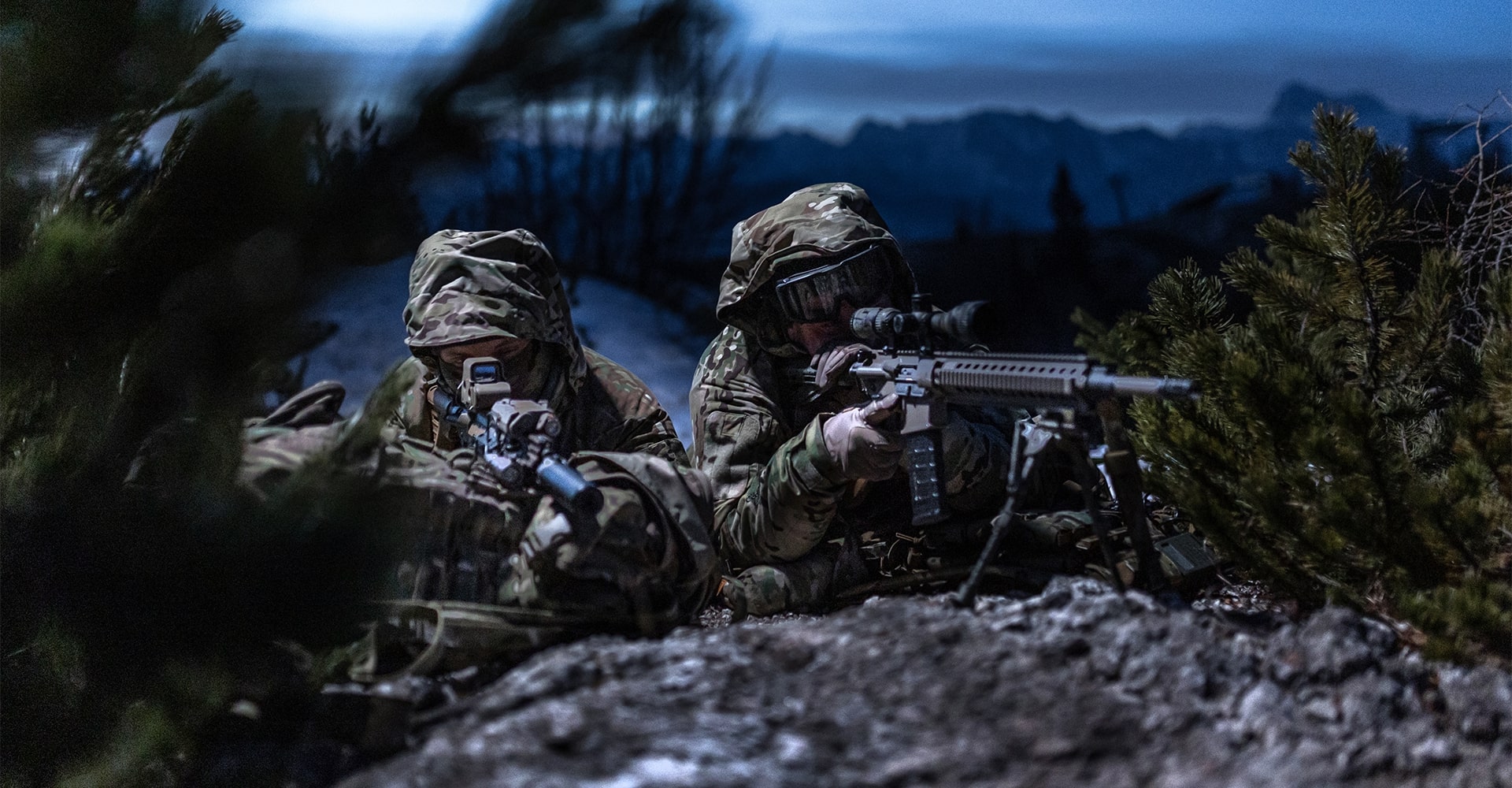 Two operators staying concealed on snowy ground in the early evening light.