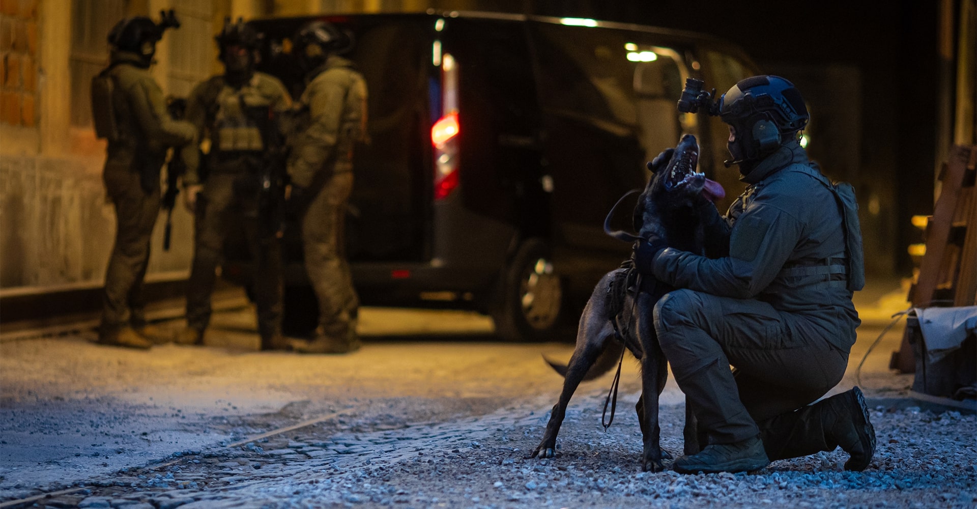 K9 dog handler kneeling and caressing his dog, while operators discuss in the background in a relaxed atmosphere.