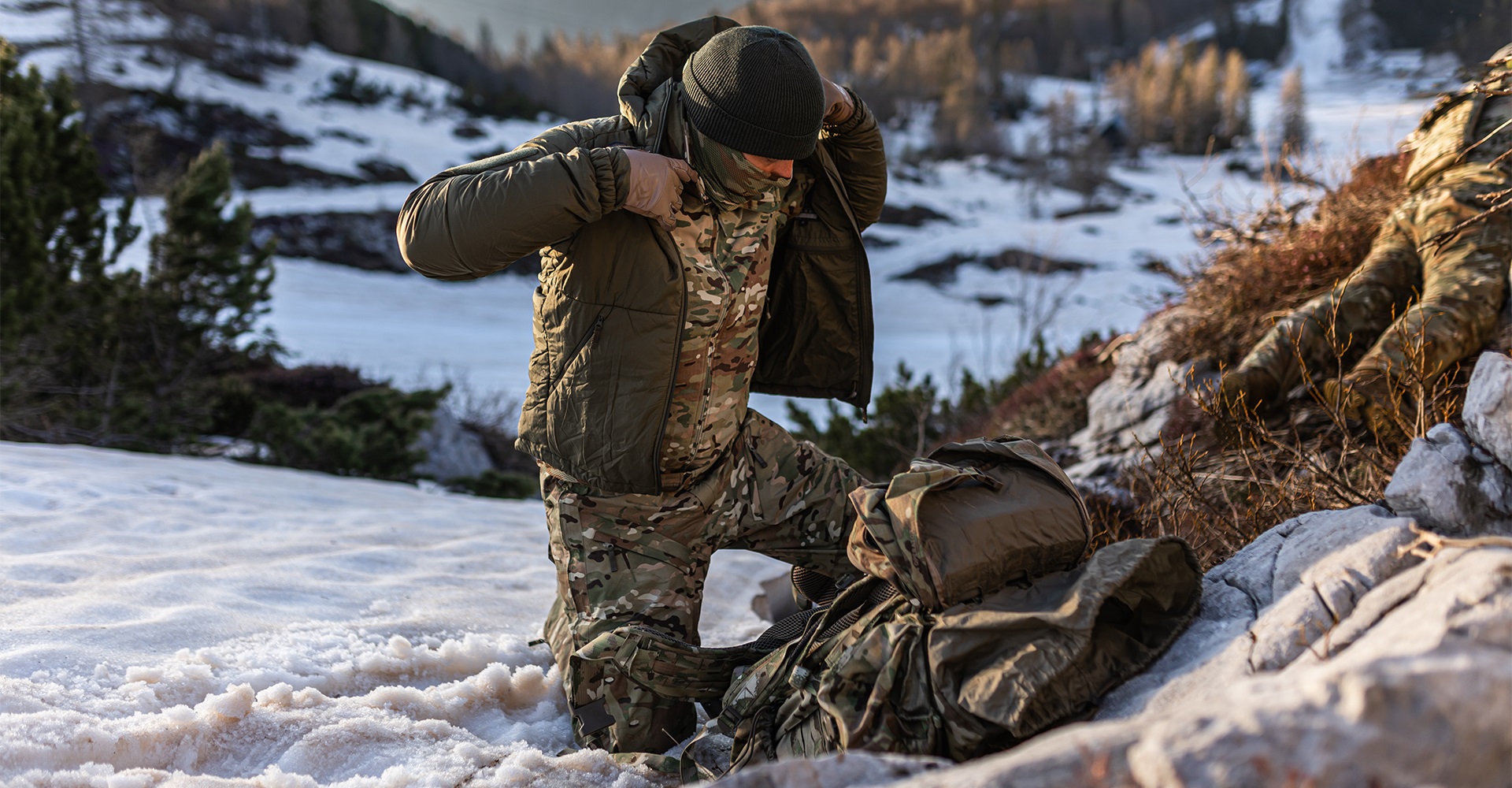 Operator kneeling on the snow, adding another layer of a tactical jacket to stay warm.