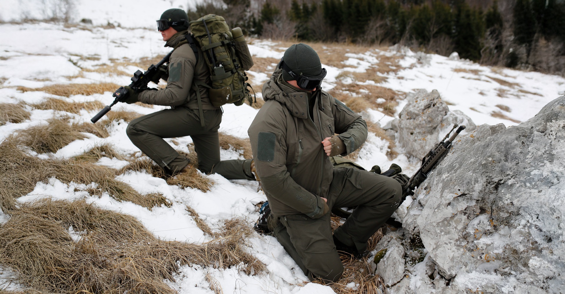 Two operatives in winter tactical clothing on a snowy terrain during a mission, one in position while the other zips up his insulated winter jacket.