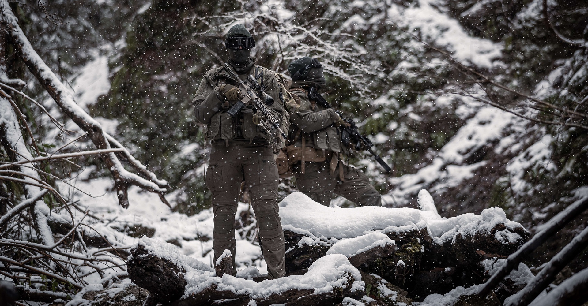 Two operators standing in a snow-covered winter forest, demonstrating the weatherproof capabilities of their tactical gear as snow falls around them.