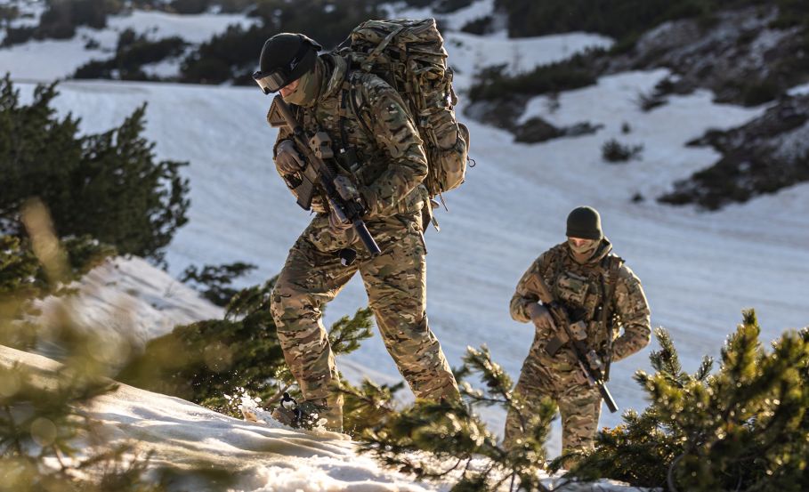 Two fully equipped operators in winter tactical clothing ascending a snowy hillside on a sunny winter day.