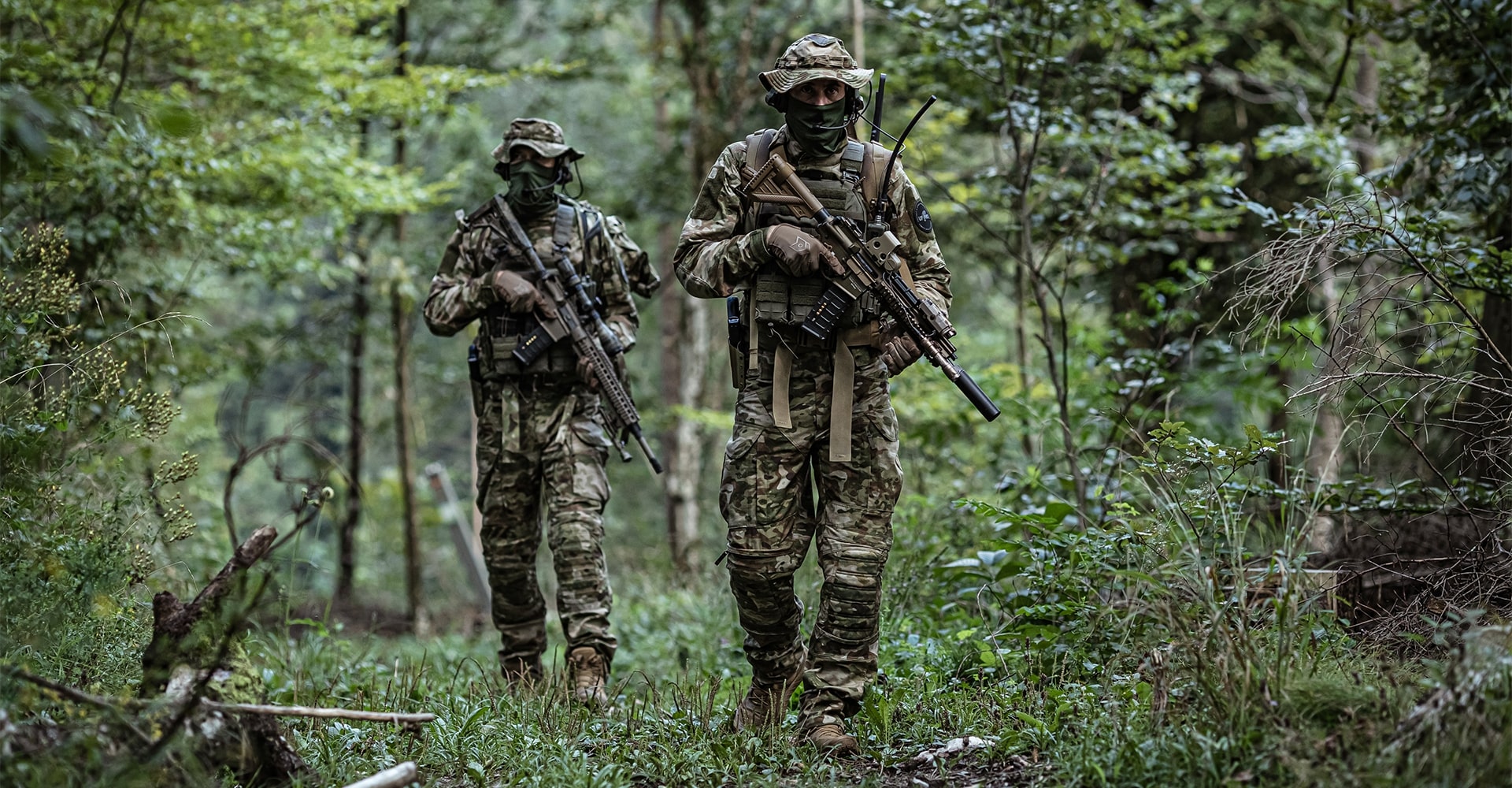 Two operators in combat gear, armed with firearms, moving through dense green forest.