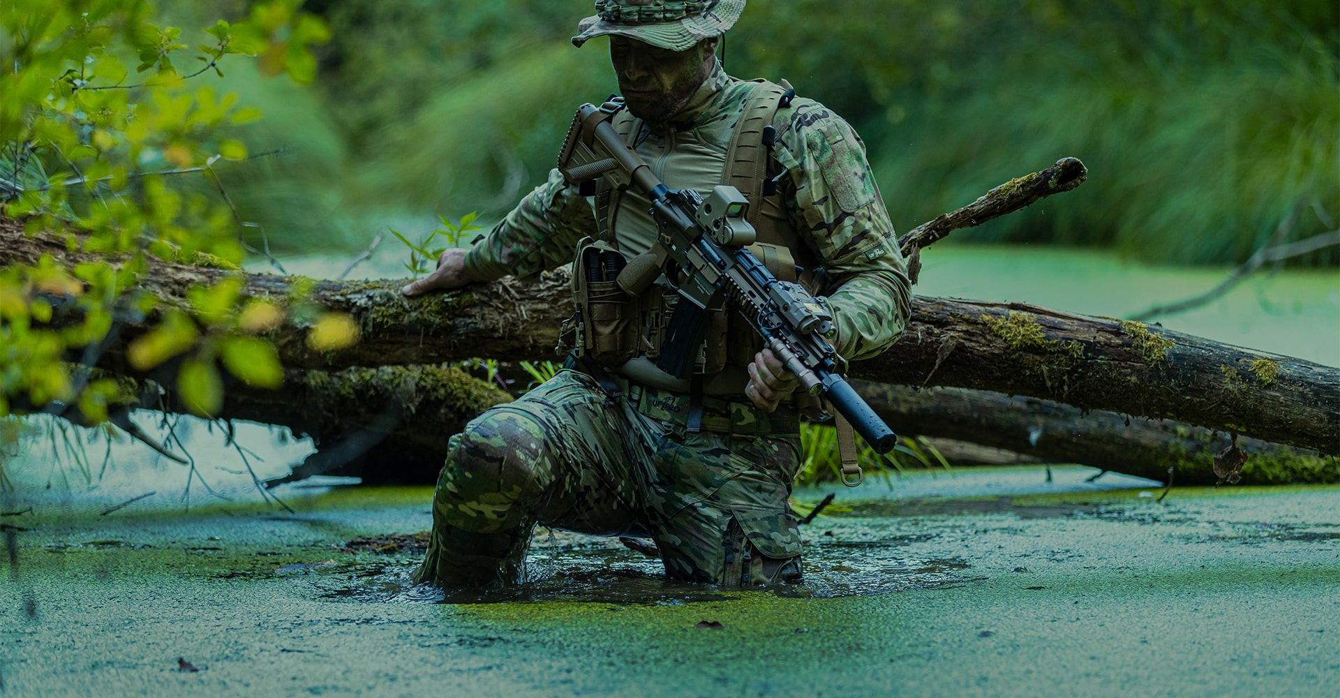 Operator in camouflage combat gear standing in muddy water in a jungle environment.