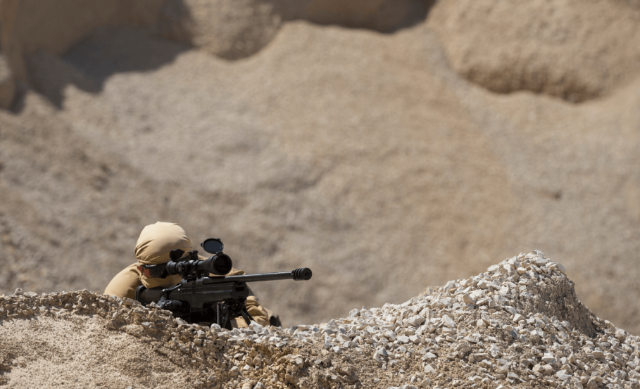 A person in beige tactical gear aims a sniper rifle in a desert-like environment, blending into the arid surroundings for camouflage.