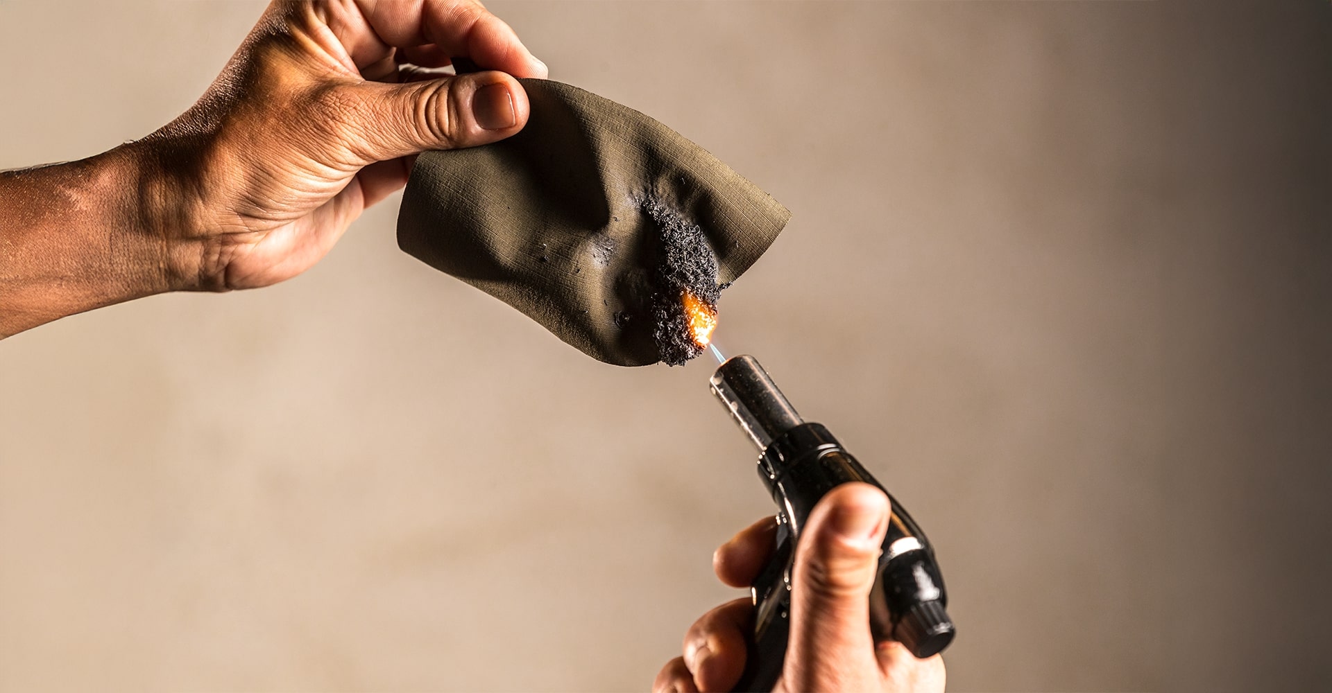 Holding a fiber cloth in one hand while igniting one end with a firestarter to demonstrate the flame-resistant properties and structure of the material.