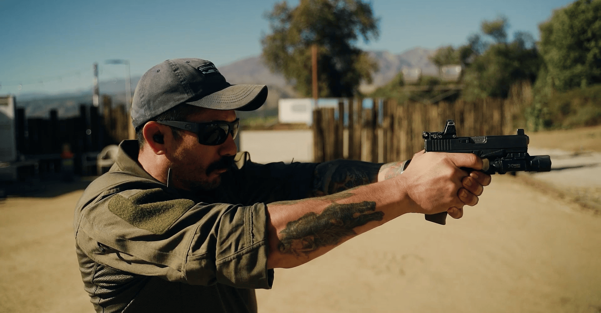 Man wearing a baseball cap and sunglasses aiming a handgun in a tactical stance.