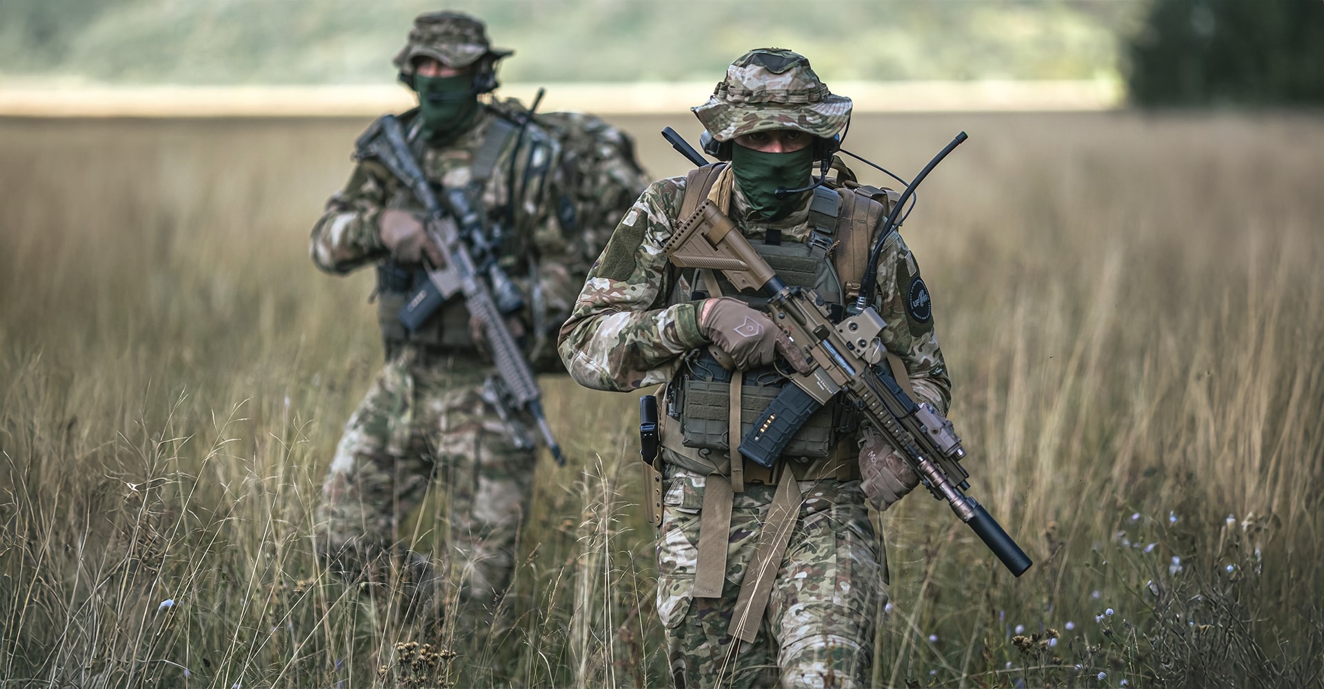 Two operators walking through tall grass, fully dressed in military BDUs.