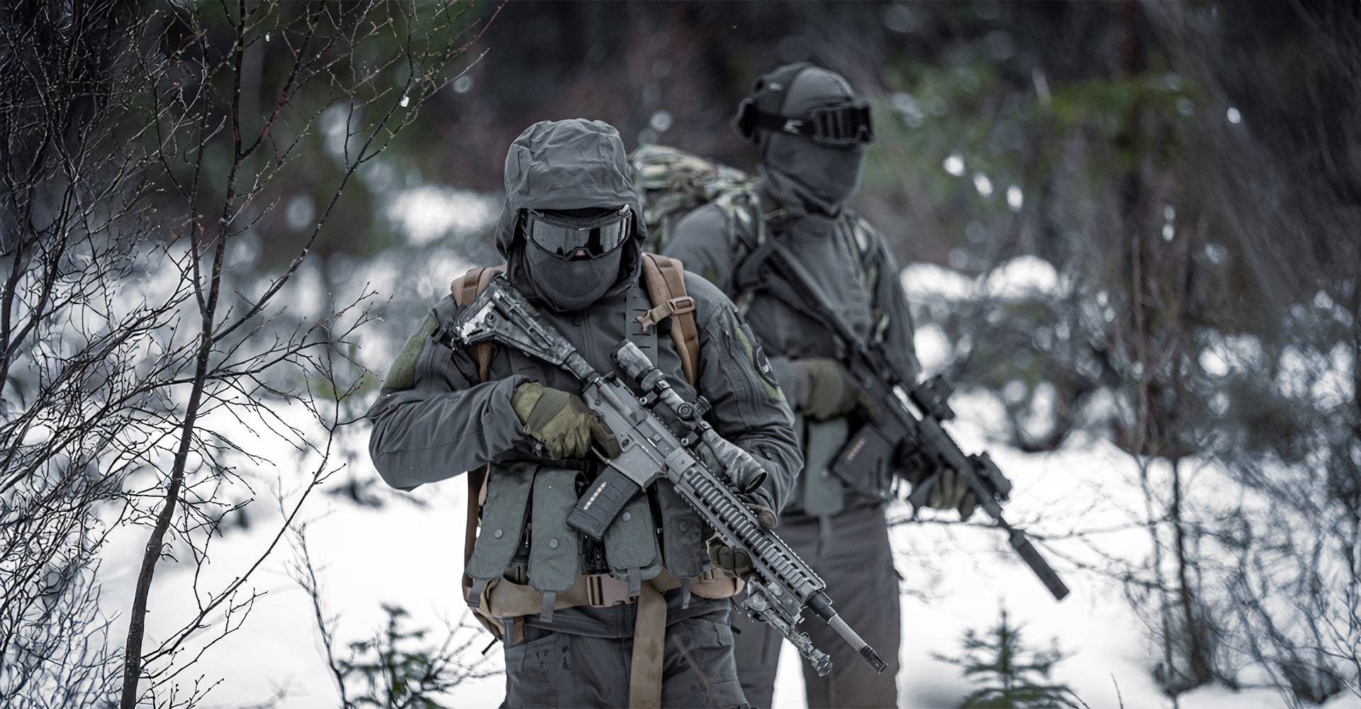 Two special forces operators walking through snowy forest terrain in full military BDUs.