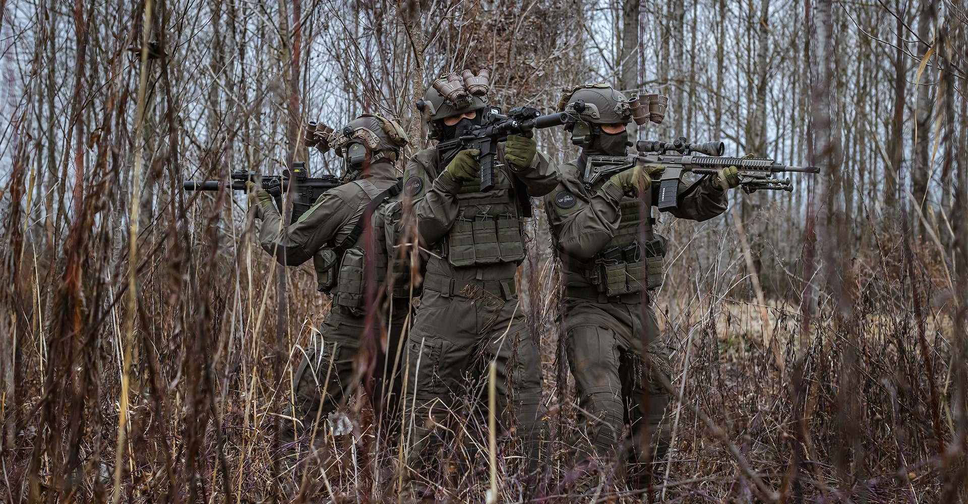 Three operators standing back to back, aiming their firearms and scanning the surroundings in late autumn European woods.