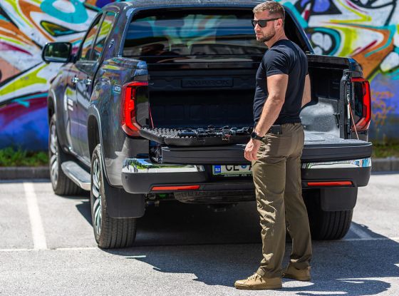 A man standing near an open truck bed in a parking lot, scanning his surroundings attentively.