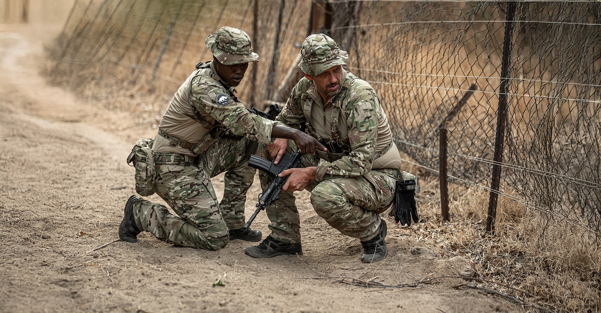 Two trackers kneeling by a dirt road in an African environment, with one pointing in a direction and the other looking where he pointed as they discuss the tracks.