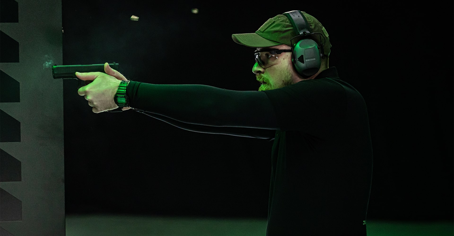 A sports shooter firing a pistol at a target, with spent bullet casings flying through the air around him.