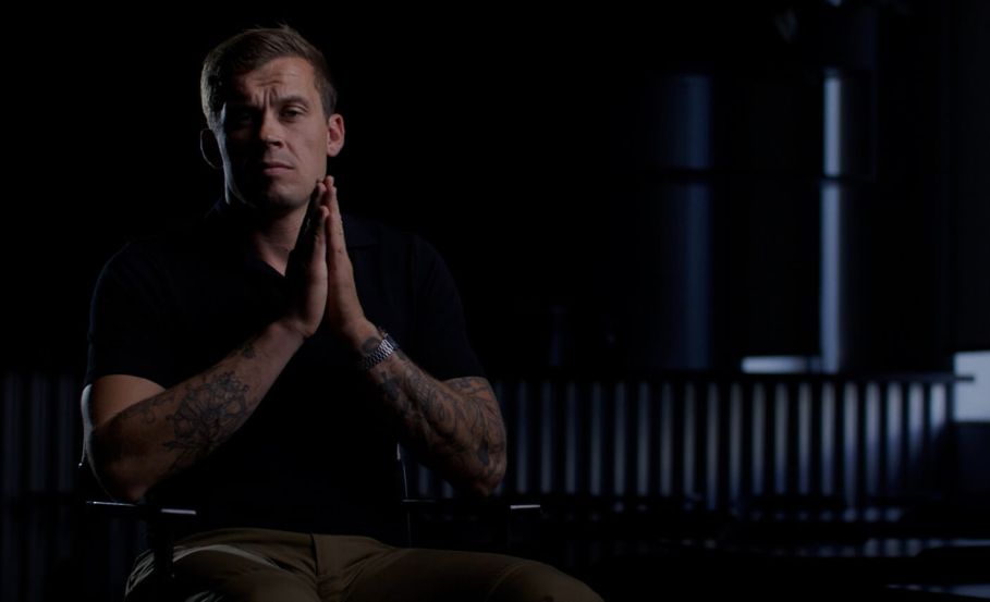 Man seated in a dark room with hands clasped, appearing reflective and focused, symbolizing contemplation of situational awareness.