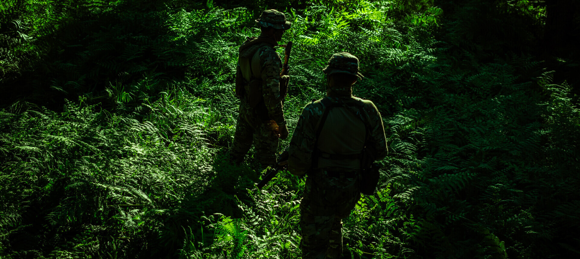 Two soldiers patrolling a dense, shadowy forest, showcasing dark-toned camouflage suited for low-light jungle environments.