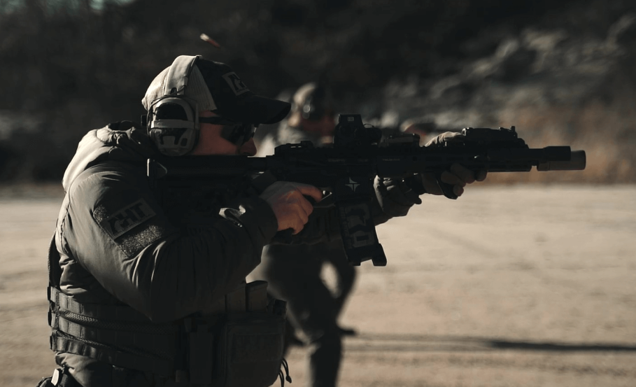 A person wearing tactical gear and headphones aims a rifle outdoors on a dirt surface with another similarly dressed individual visible in the background.