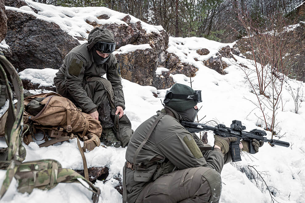 Two tactical operators taking cover behind a rock and one putting on Delta OL 4.0 winter pants