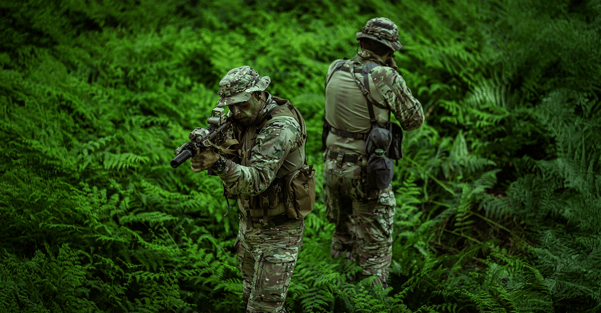 Two operators in dense jungle terrain wearing full camouflage battle dress uniforms and tactical gear, carefully surveying their surroundings for potential threats.