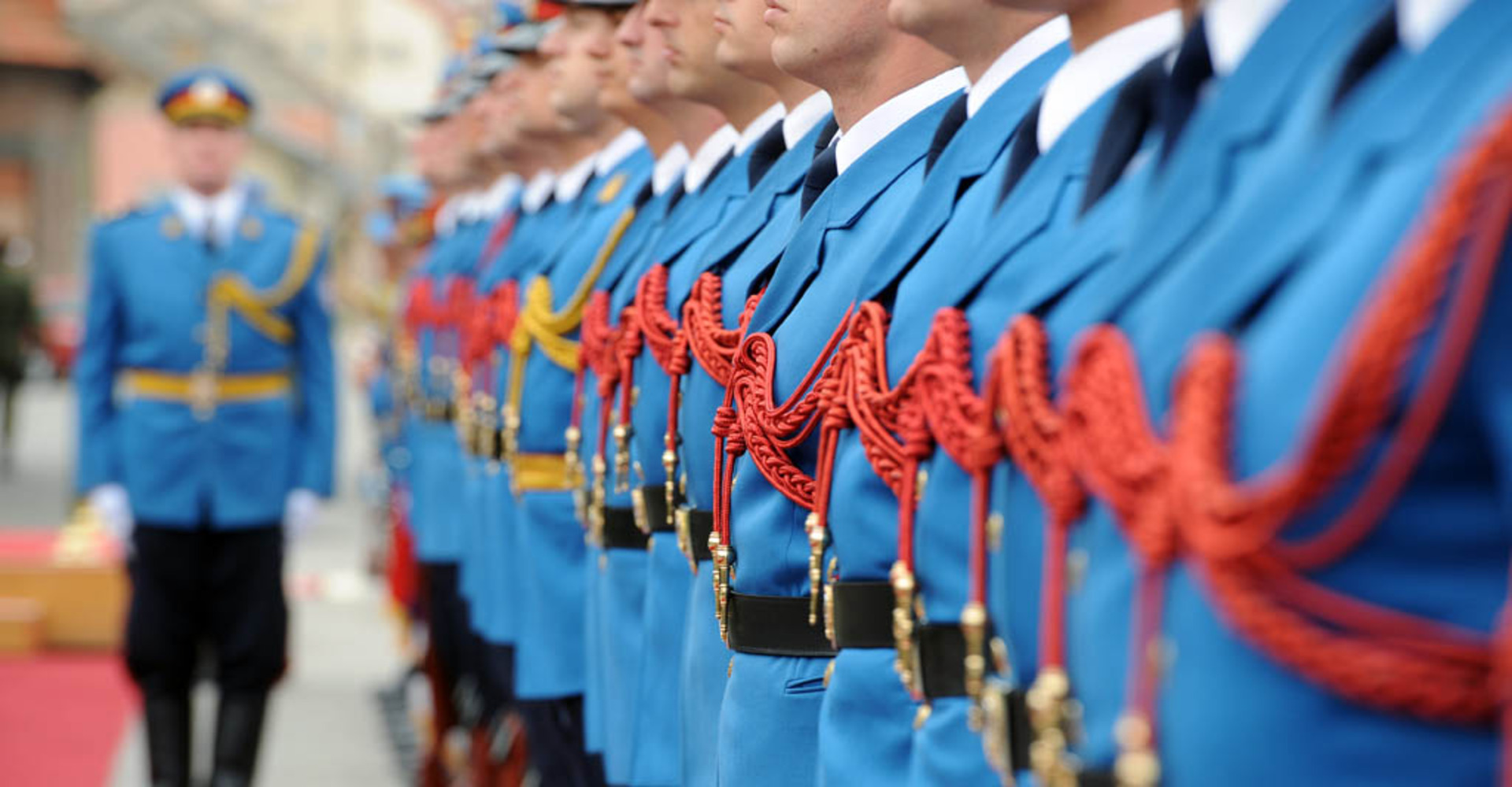 Men standing in a formal line during what appears to be a ceremonial event, dressed in bright red uniforms with gold accents, maintaining strict posture.