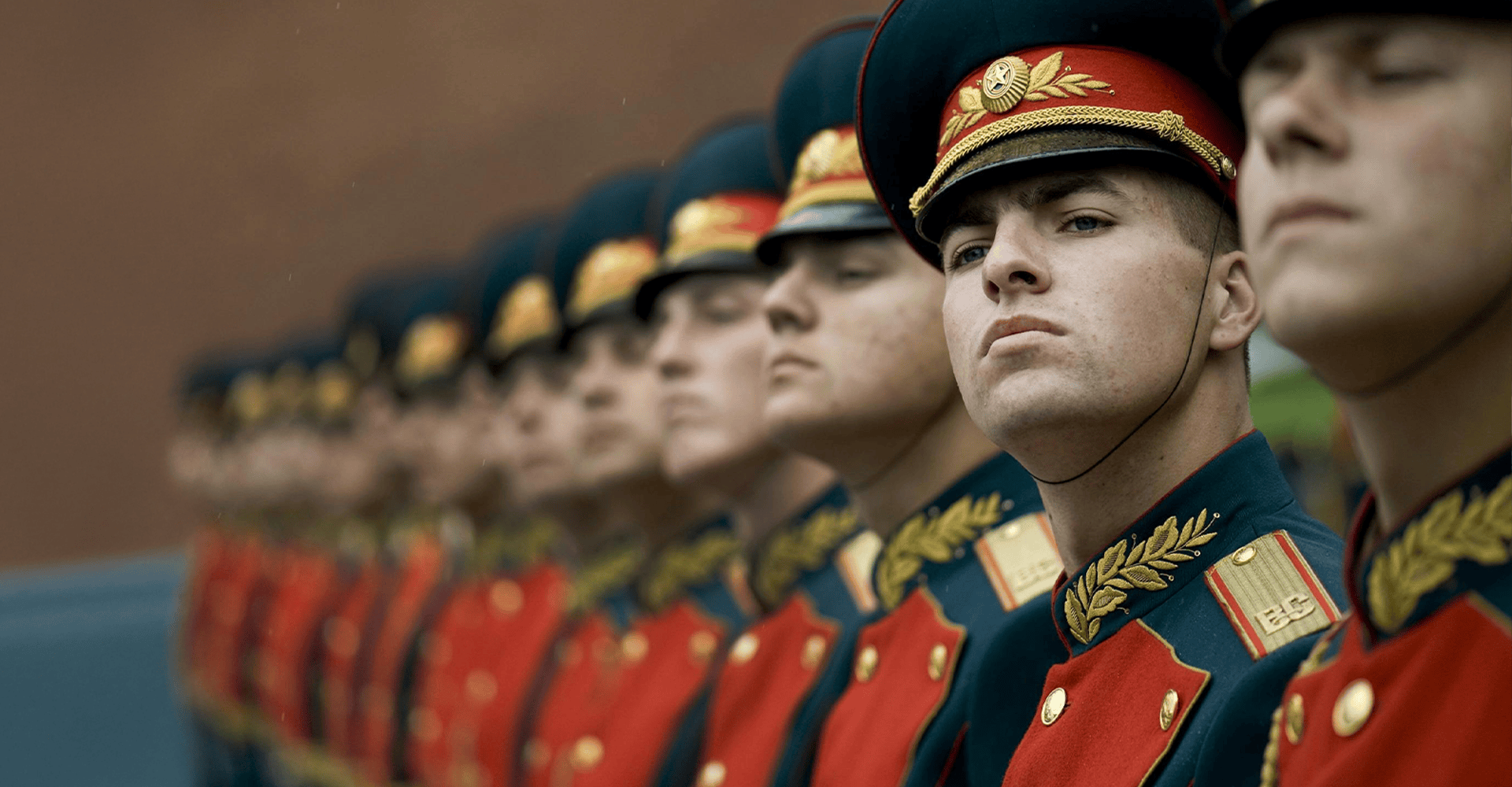 Men standing in a formal line during what appears to be a ceremonial event, dressed in bright red uniforms with gold accents, maintaining strict posture.