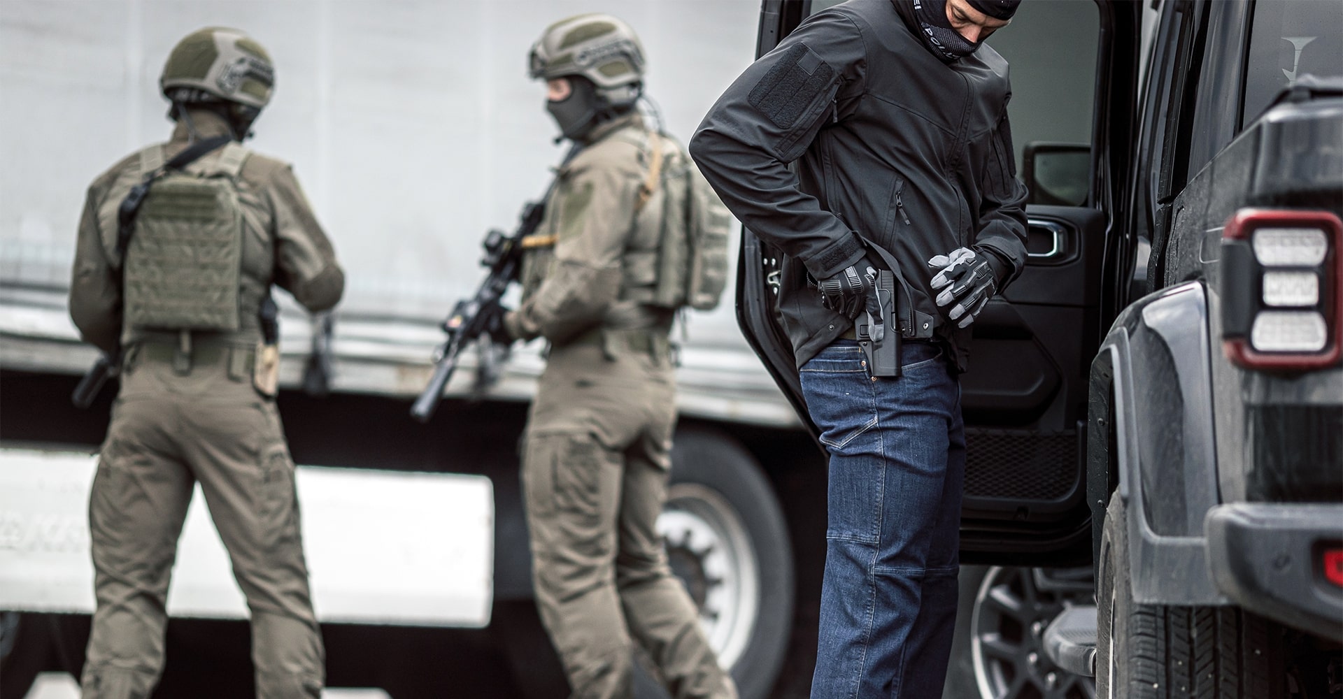 A police special forces operator getting ready in a civilian environment, with cars and trucks in the background, while two operators stand guard.