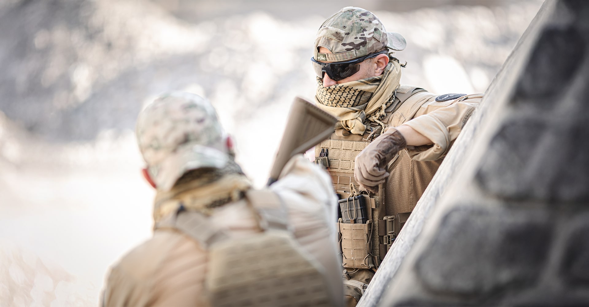Two operators in a military setting, likely in a hot environment, as indicated by their tan-colored uniforms and the rocky background, taking a break in between a mission.