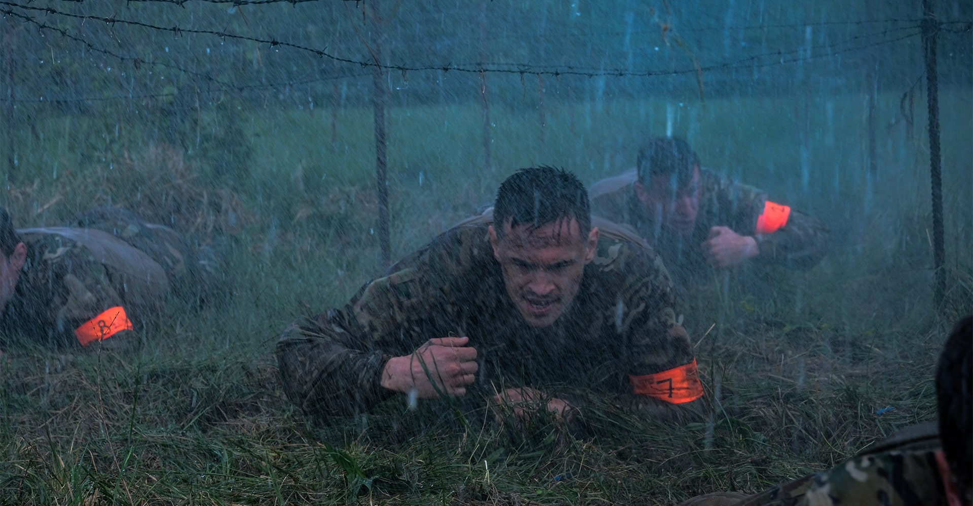 Special forces selection process with three attendees crawling under barbed wire in heavy rain.