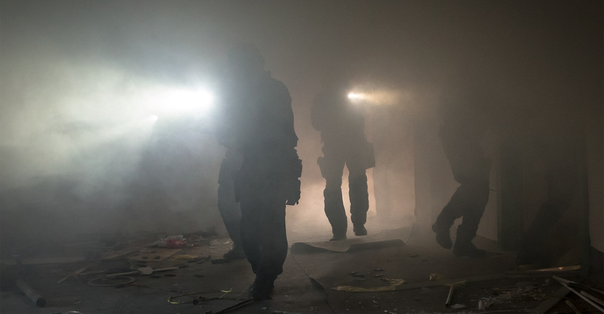 Operators entering a smoke-filled room, using their firearm lights to illuminate the debris-littered space.