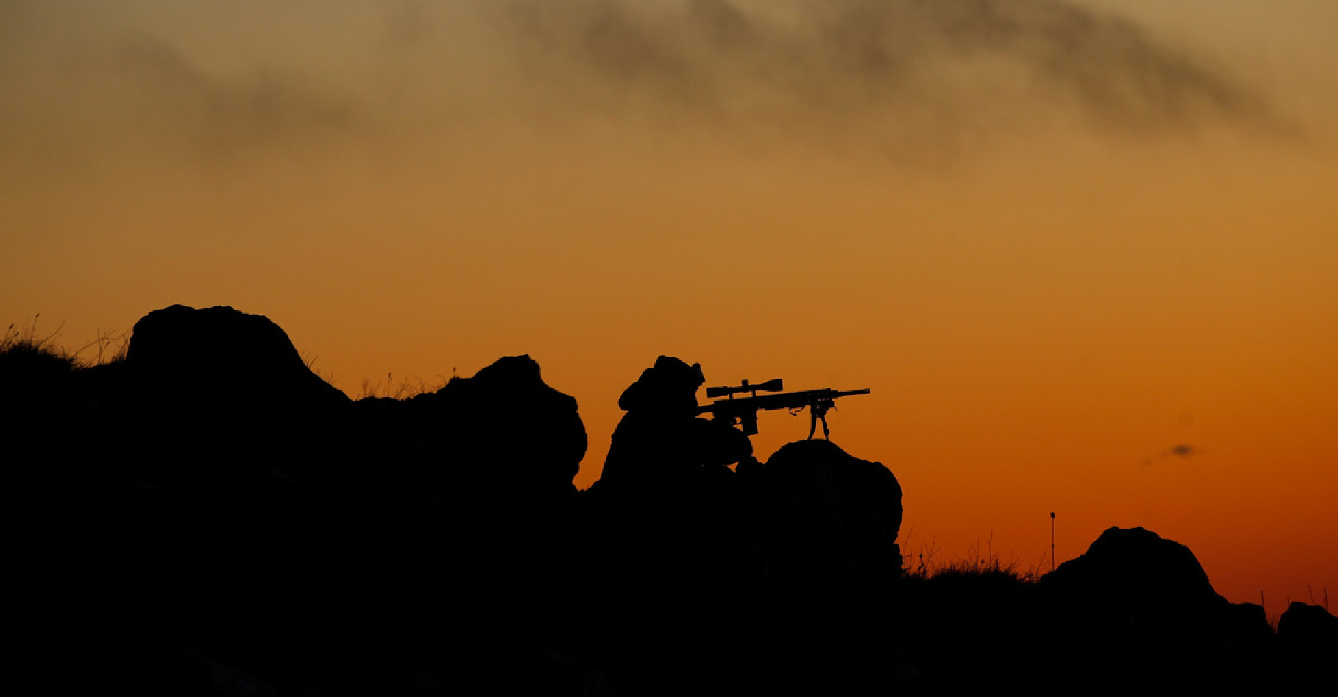 A silhouette of a man with a firearm, positioned behind rocks at sunset.