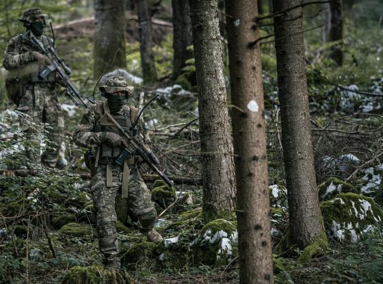 Two operators moving cautiously through the woods, blending into their surroundings with camouflage clothing.