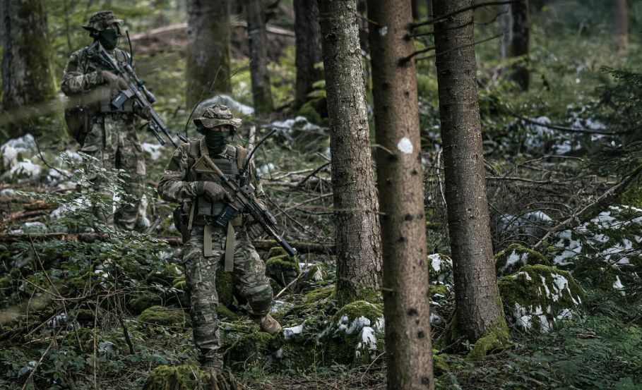 Two operators moving cautiously through the woods, blending into their surroundings with camouflage clothing.