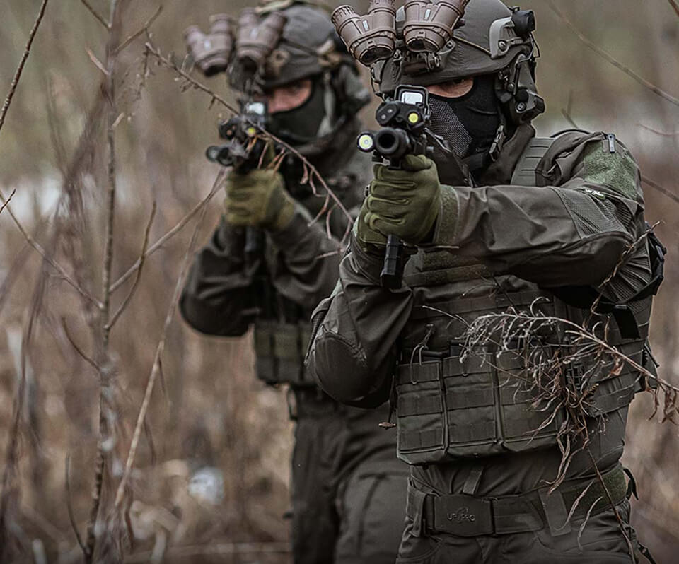 Two tactical operators standing in the underbrush wearing Striker XT Gen.3 Combat Shirt and carrying a rifle.