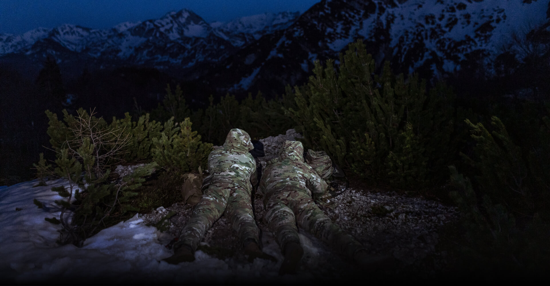 MISSION-SPECIFIC COLD WEATHER CLOTHING Two soldiers in MultiCam winter gear lying on a snow-covered hill, with mountains in the backdrop.