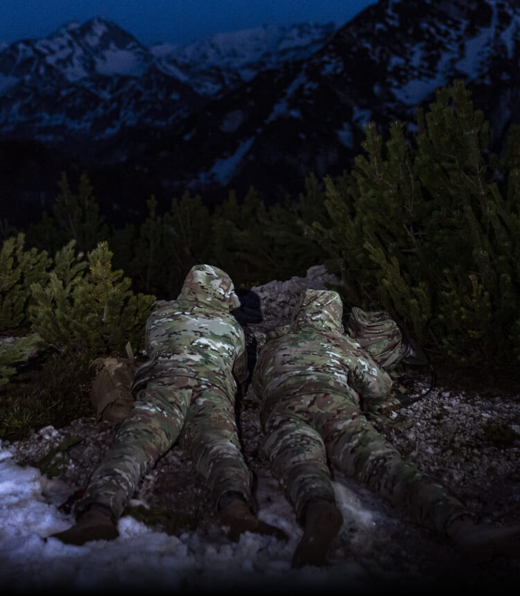MISSION-SPECIFIC COLD WEATHER CLOTHING Two soldiers in MultiCam winter gear lying on a snow-covered hill, with mountains in the backdrop.