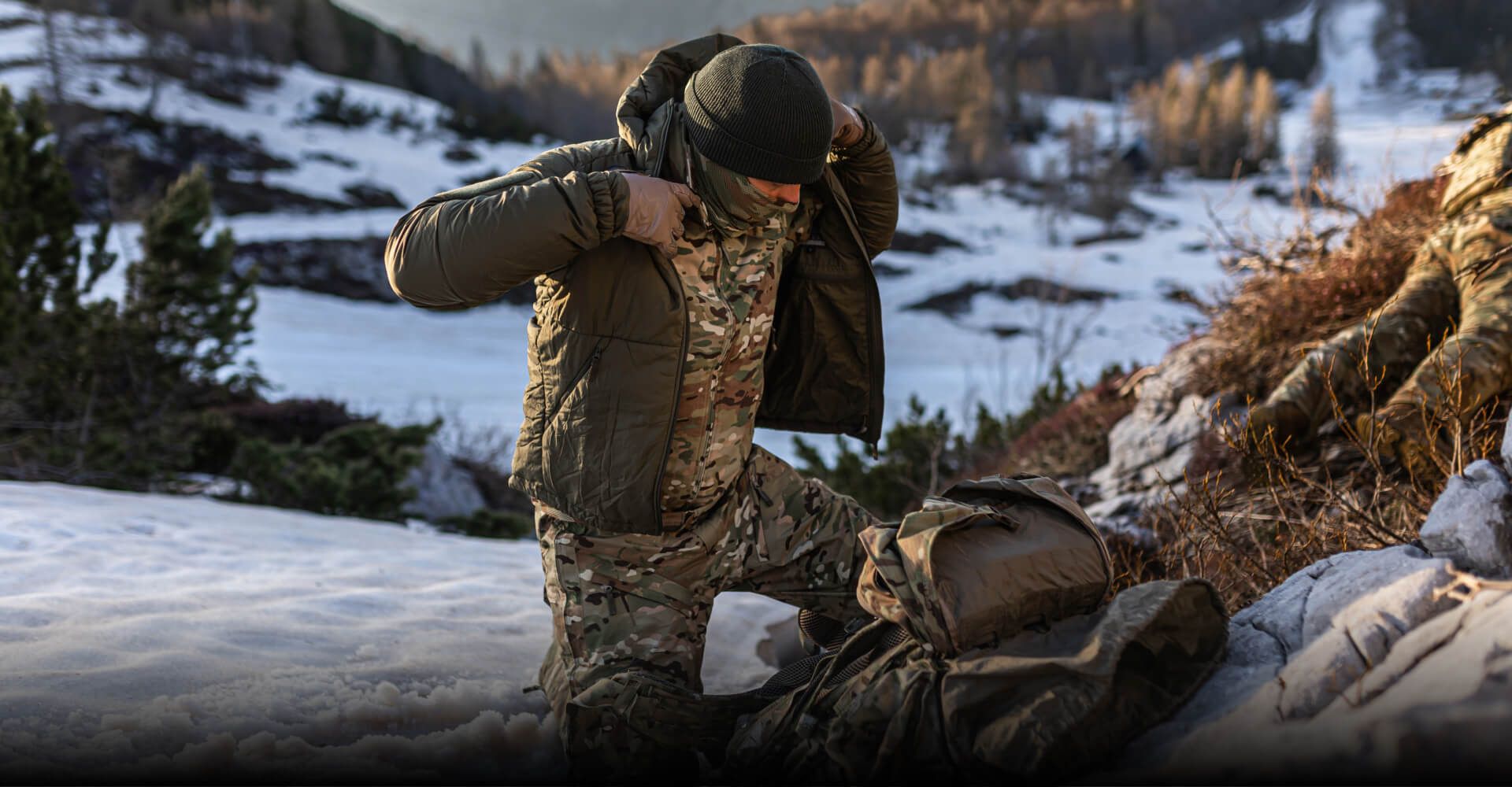 LAYERING SYSTEMS Alt text: Soldier putting on an insulated tactical jacket over MultiCam winter pants and jacket in snowy mountain terrain.