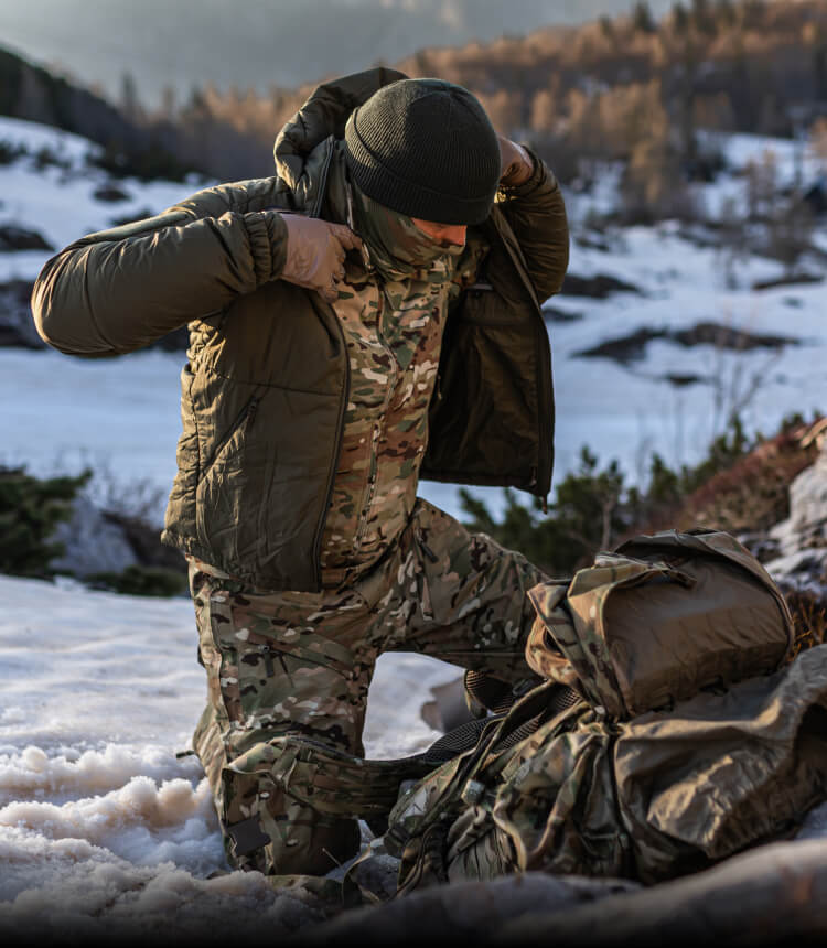 LAYERING SYSTEMS Alt text: Soldier putting on an insulated tactical jacket over MultiCam winter pants and jacket in snowy mountain terrain.
